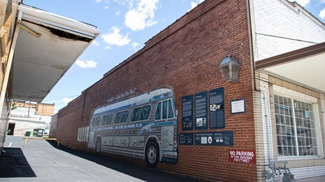 Painting of Greyhound bus and panels on a brick wall on alley side of a storefront