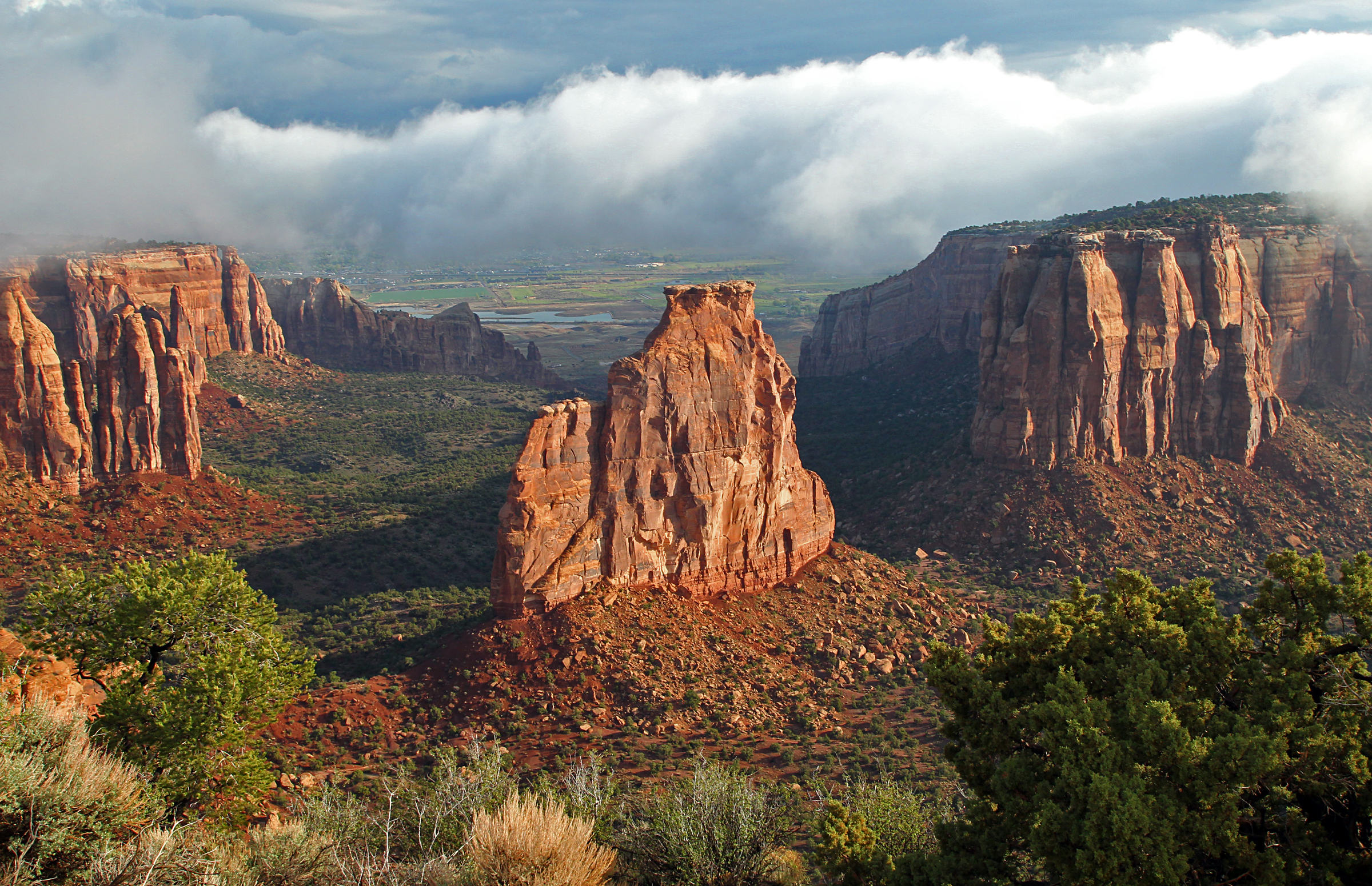 reddish sandstone rock tower standing above green bottoms with cliffs and clouds around