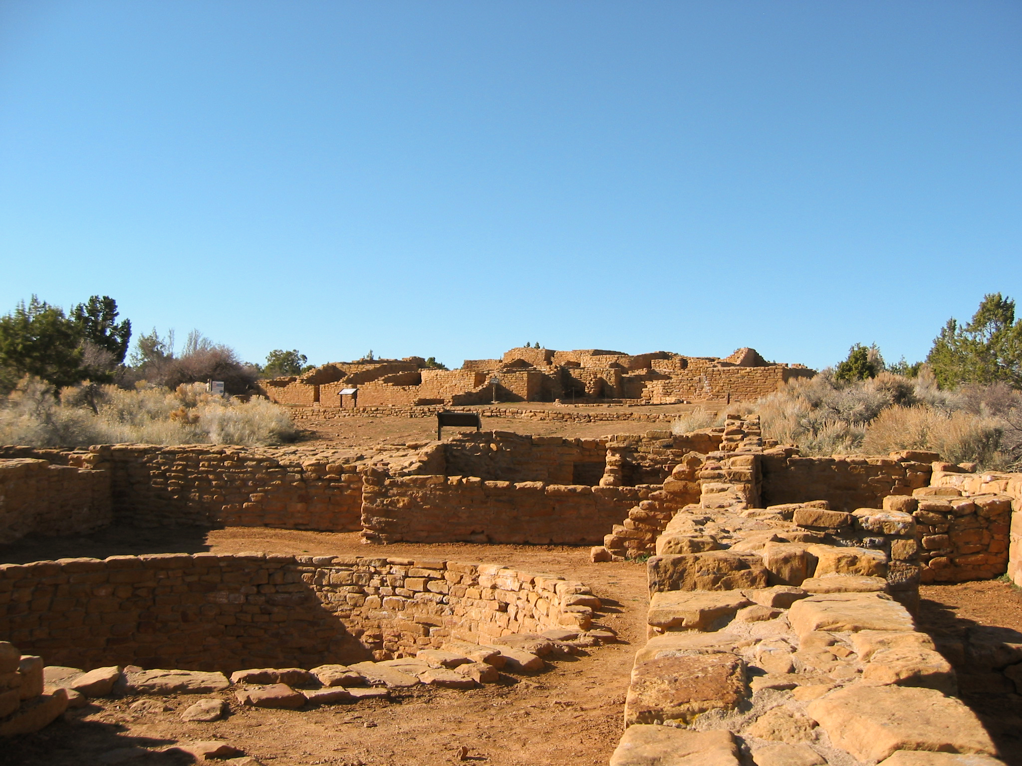 View across low, stone-masonry walls, both square and round. In the background is a one- and two-story remains of a multi-roomed pueblo.