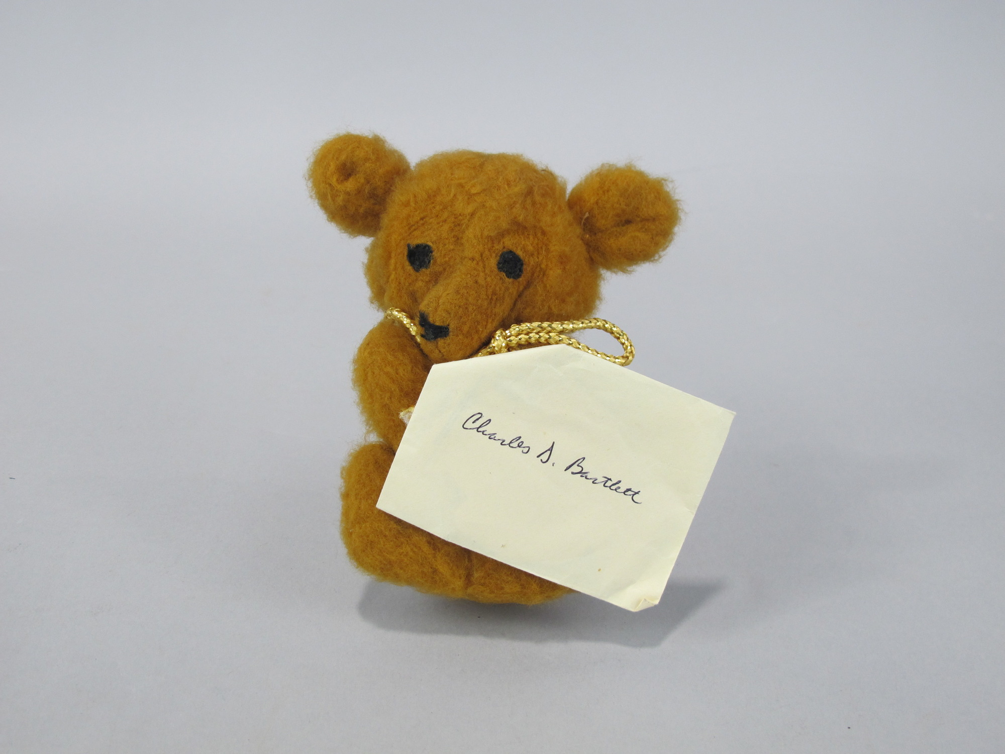 Small stuffed bear with a card attached to a gold string tied around its neck. The card's envelope is addressed to Charles Dennis Bartlett (KIA 08/08/1968). The card inside is in the shape of a bear and  says "To: Denny From: Your cousin, the Old Bear still remembers you".
