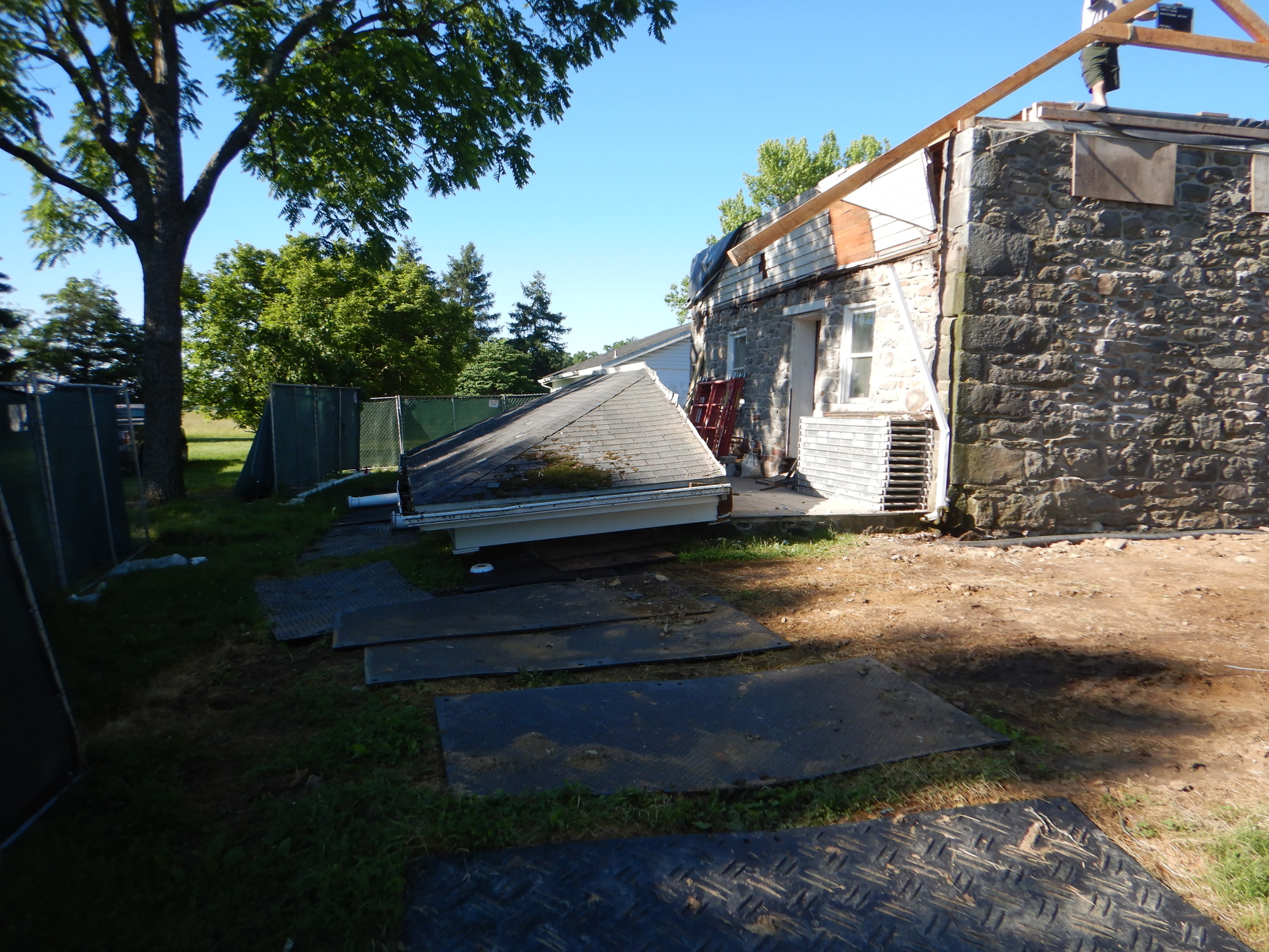 The photo is taken outside. The green construction fence is still up. There is a worker on the flat roof. The porch roof is intact but is on the ground. It is no longer attached to the house.