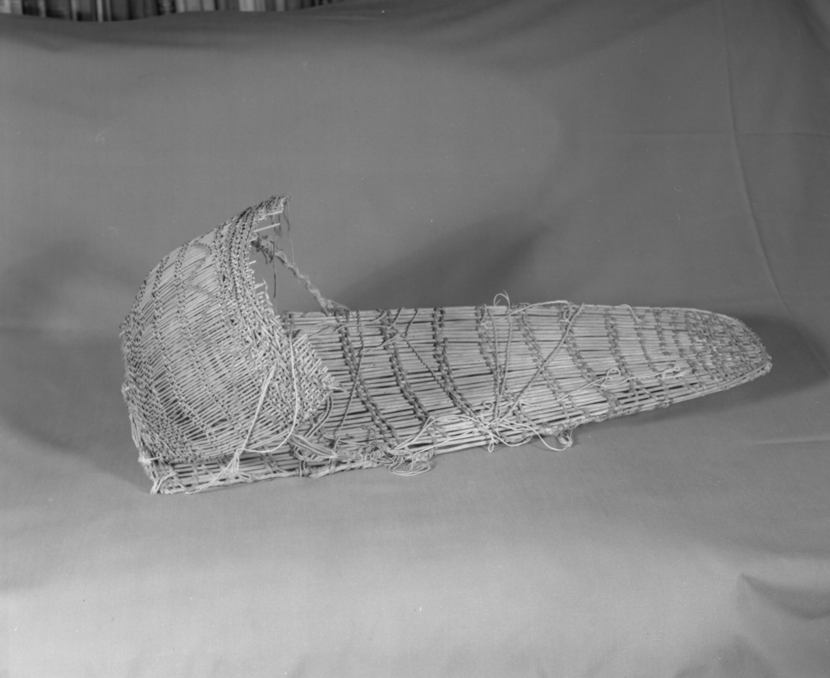 Private Collection. Object: Miwok cradle basket. Dimensions: 26 x 8 1/4. Hood 10 x 12". Bow projections: 6 1/2". Materials: Split bull pine, willow, red floss, white cotton cordage. Technology: Plain twined, up to right slant of turns, warp at start are trimmed and bent over another rod and plain twined. Strengthening devices as on previous cradle ends of cross rods are trimmed and stitched to side of cradle. Hood has paired rows of twining slanting in opposite directions below bow as well as four rows of three strand twining in hood. It is unusual in that there are fairly closely twined sections below the bow, and one near the back selvage. Collection History: Made by Matilda, a Sonora Miwok, c. 1920. Photographed by Michael Dixon. Catalogued by Craig Bates and Barbara Beroza. Yosemite Collections, National Park Service.