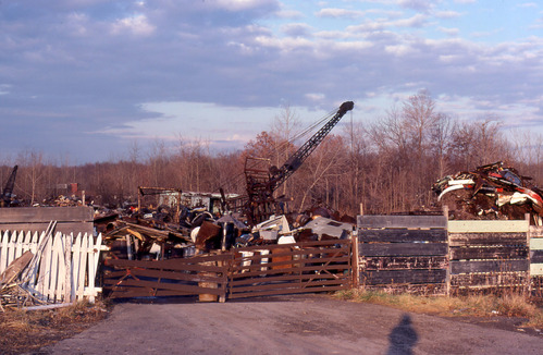 Behind a fence of mismatched wood and metal is a large industrial waste dump. A crane and pulley stretch to the sky and bare trees are in the background. 