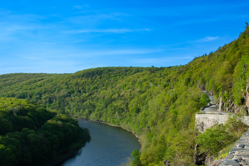 View of lush, green rolling mountains with blue river running through it. On the right is a two-lane winding paved road, carved into the hillside, supported by a stone wall.