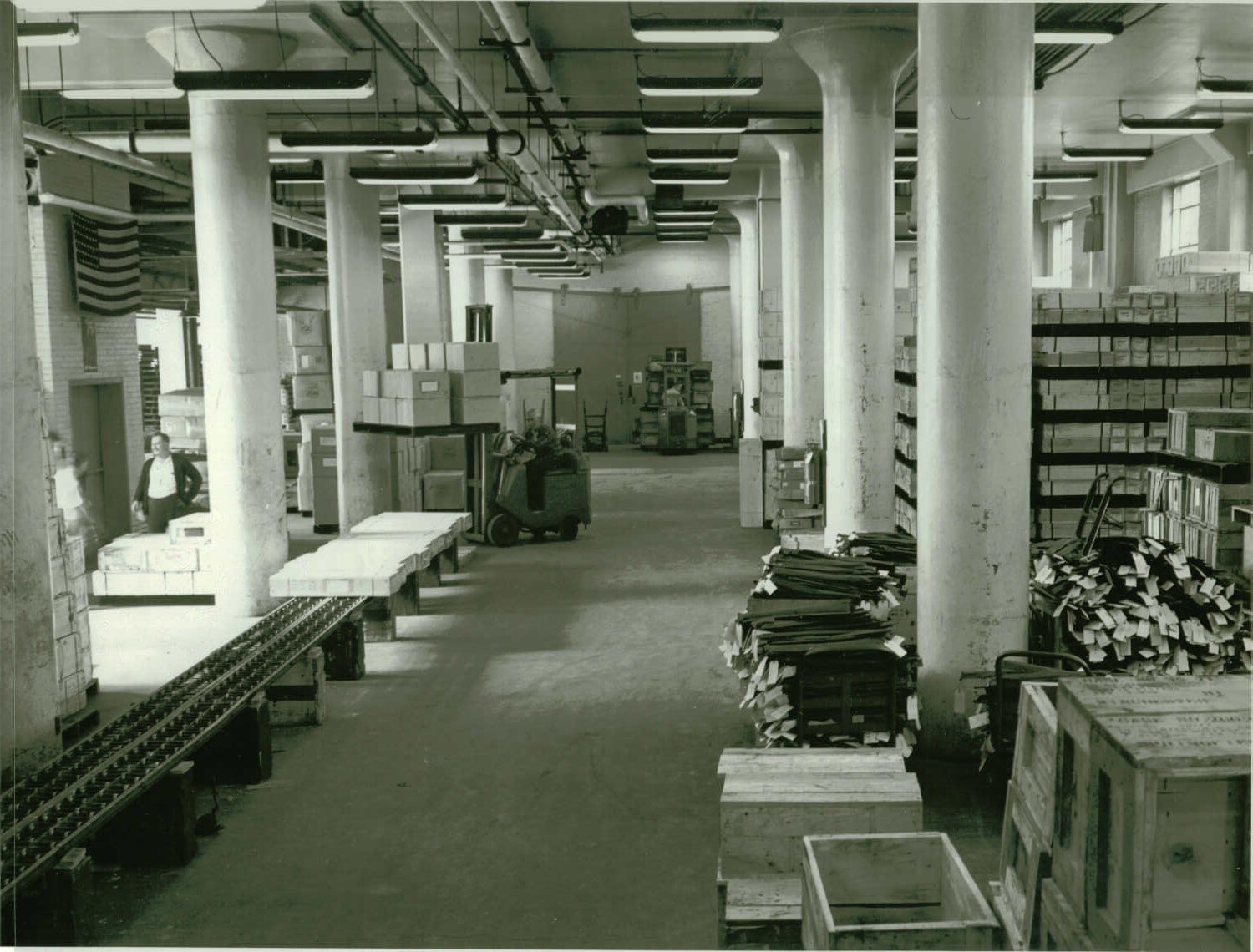 Black and white photo of a warehouse room. Pillars run along the right and left sides of the room. Boxes and carts are stacked between and behind the pillars. A roller table with long boxes at the end of it sits is along the let pillars. A man drives a forklift with a pallet full of boxes near the end of the roller table.