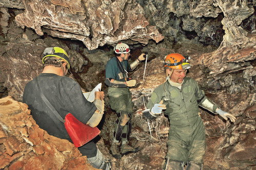 3 men in hard hats and dirt covered clothes survey a small cave passage. 