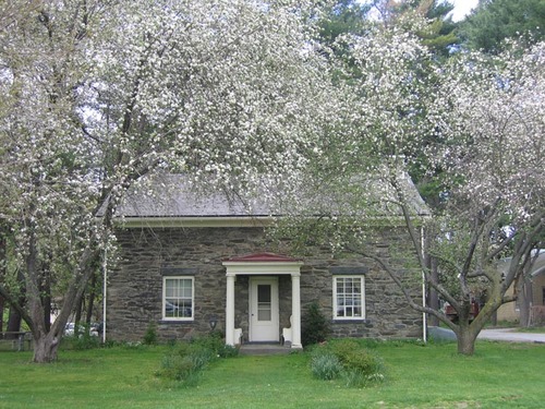 Trees blossom on each side of a two-story, field stone house with two windows opposite a door.