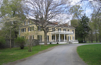 A driveway leads a large two-story mansion with a white porch, yellow siding, and black shutters.  