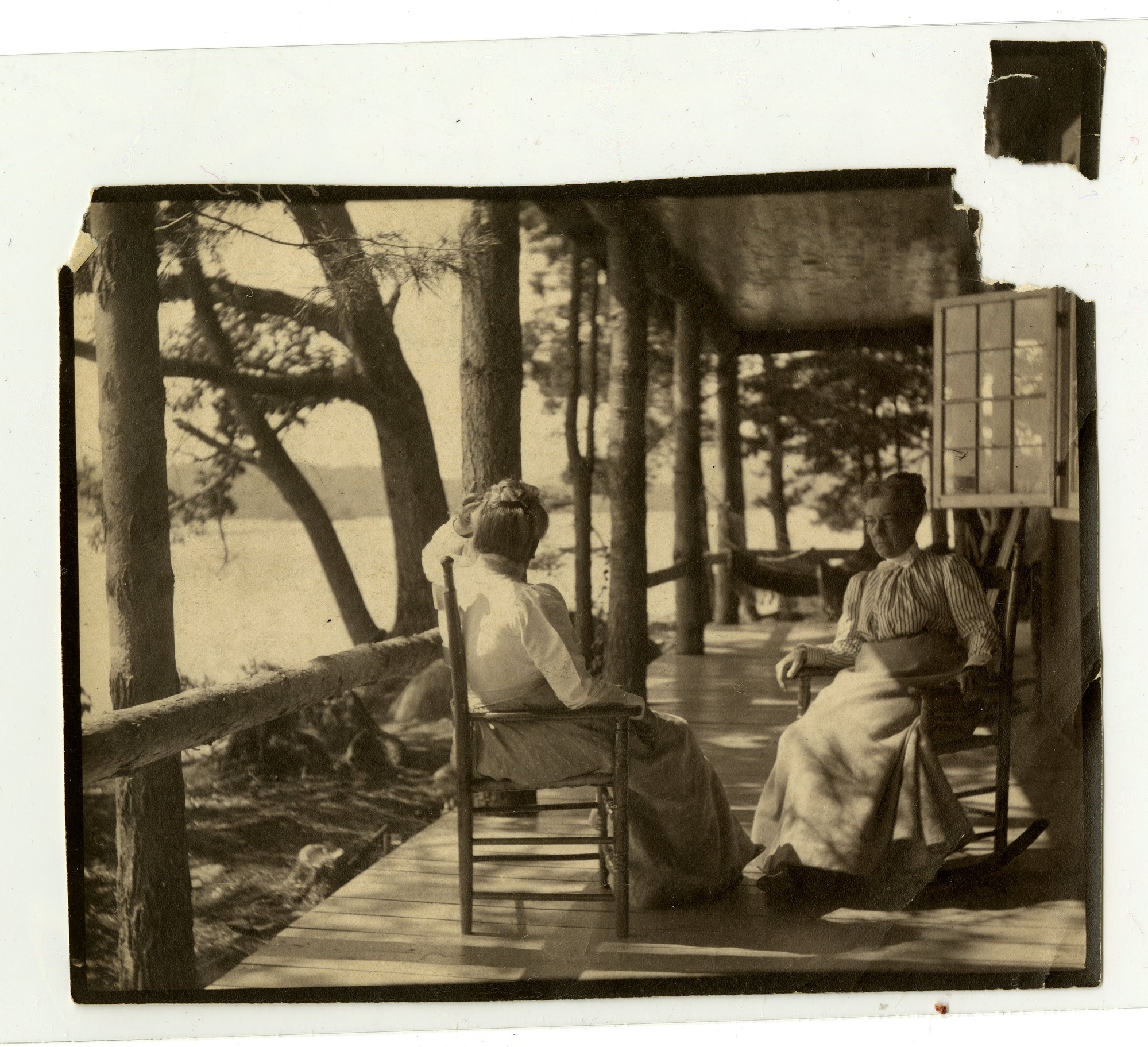 Two women seated on long rustic porch, looking over lake. One woman faces towards the camera in a rocking chair, the other faces away.
