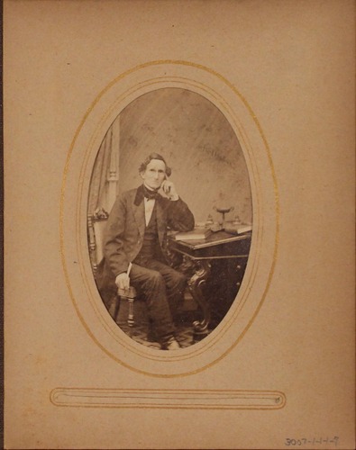 Black and white photograph of man sitting at writing desk looking at viewer.