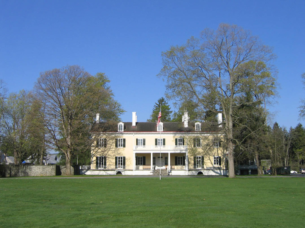 View across the lawn to the flagpole, trees, and broad two-story mansion with many windows.