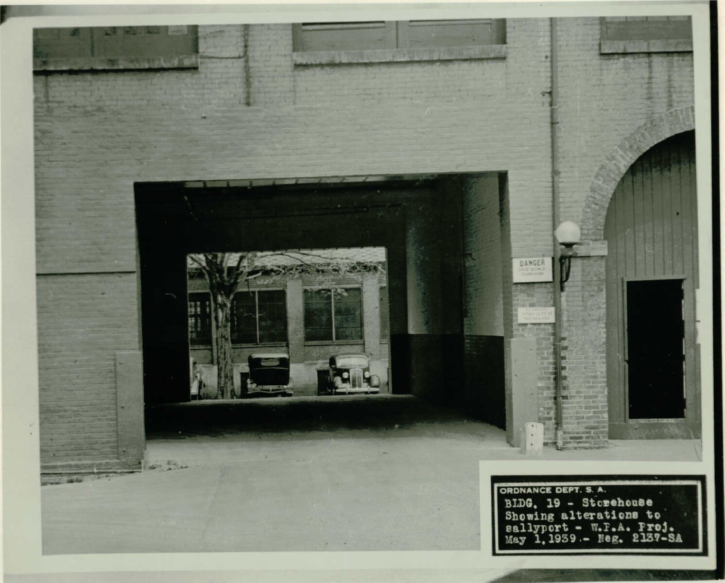 Black and white photo of an open portion through a brick building. Parked cars and a tree can be seen on the other side. Text on the photo reads, “Ordnance Dept. S.A., BLDG. 19 – Storehouse, Showing alterations to sallyport – W.P.A. Proj. May 1, 1939 – Neg. 2137-SA.”