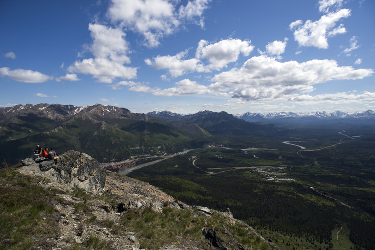 people sitting on an outcropping on a mountain, overlooking a forested valley with river and road through it