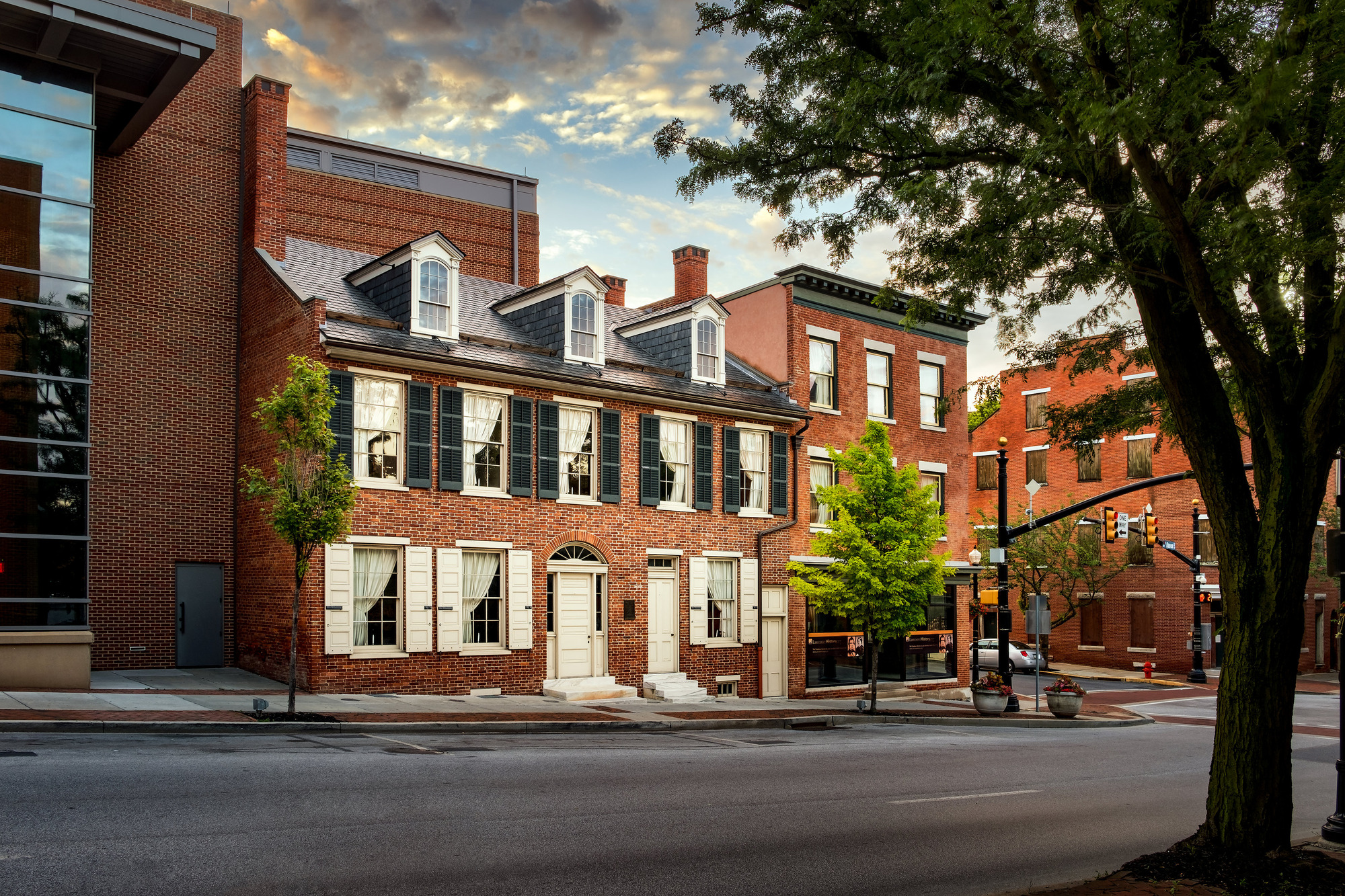 A three-story red brick colonial style house with shutters on the windows and three dormer winders in the roof. The building sits on a city street surrounded by newer red brick buildings.