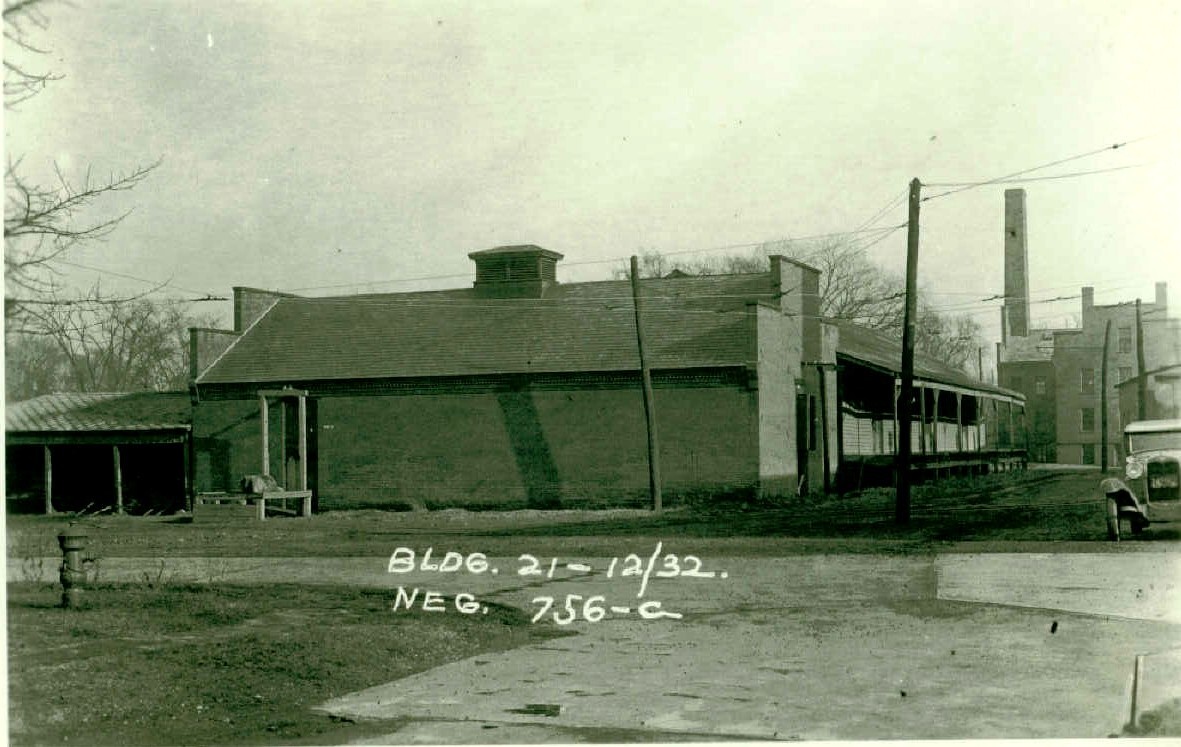 Black and white photo of a brick building with a vent in the center of the roof. An open shed is behind it. Another building with a covered porch is on the other side of the brick building. A car is parked out front. A fire hydrant is across the road in the left foreground. Writing on photo reads, “Bldg. 21 – 12/32. Neg 756-C.”