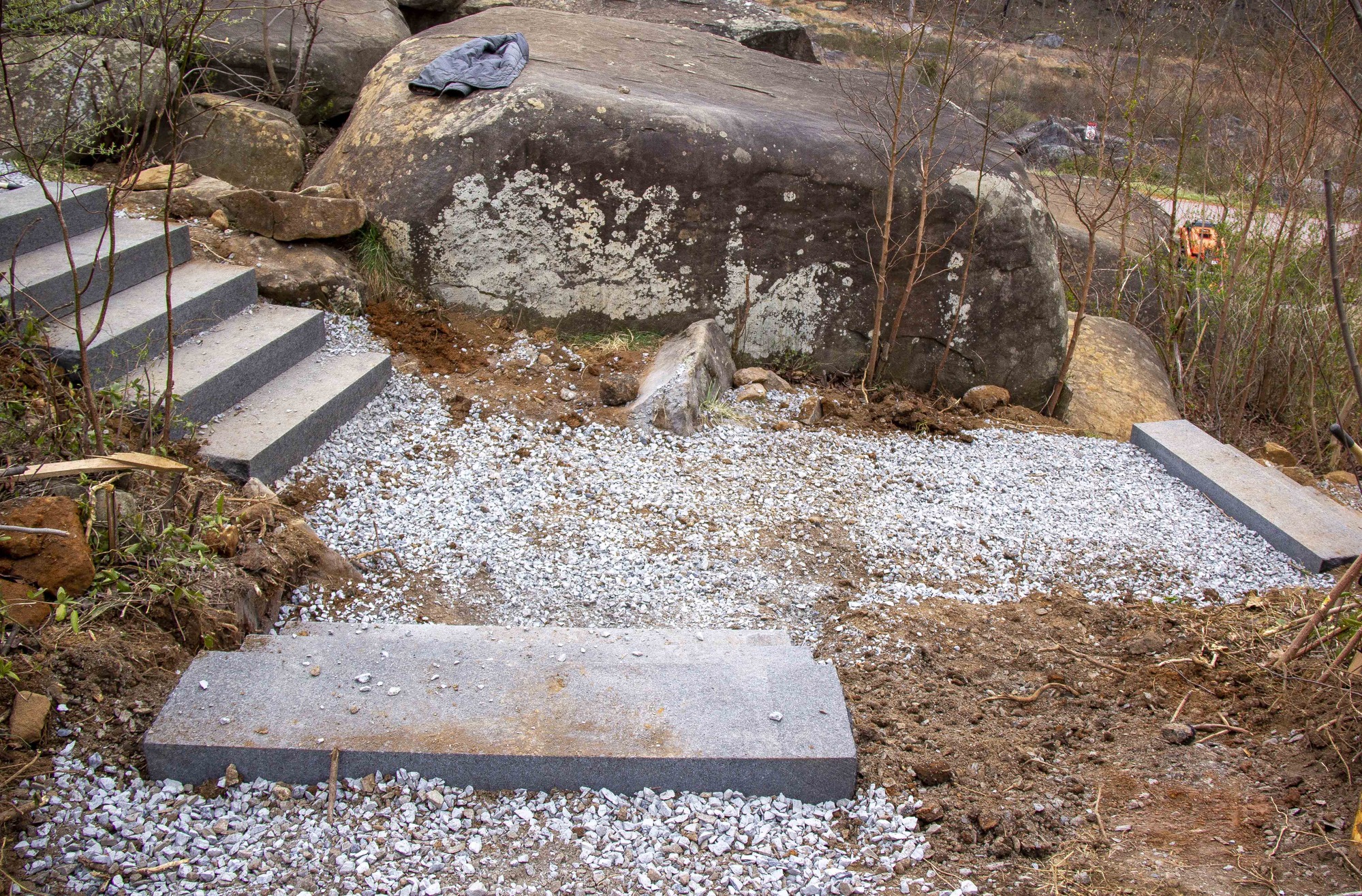 In center frame is a gravelly Y-shaped path leading down to the right, up to the left, and up towards the camera. On the left are five newly installed stone steps leading up the side of a small slope. There are three stairs leading up towards the camera and other leading down off the right of the frame.