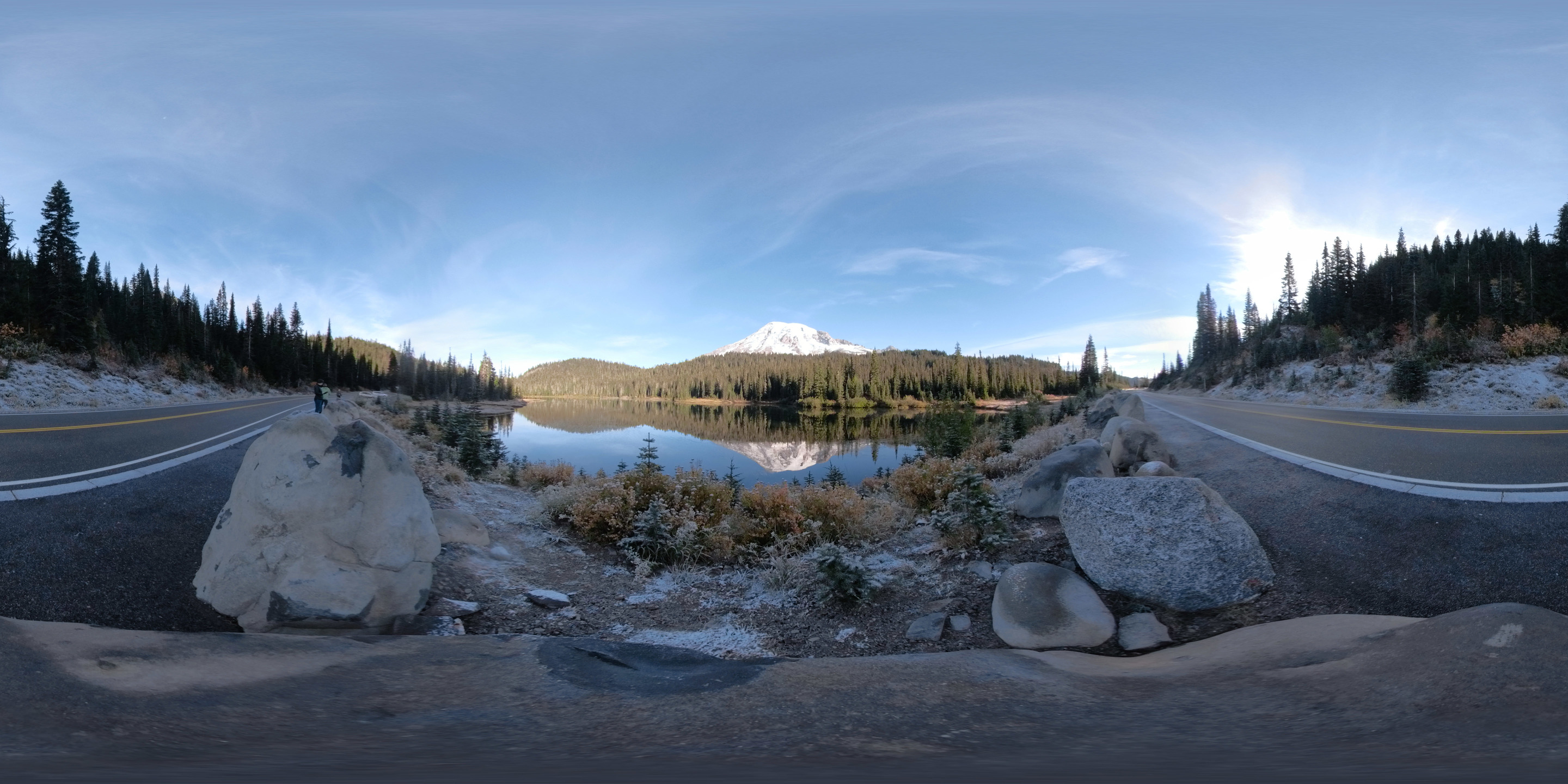A glaciated mountain surrounded by forested hills reflects in the still waters of a mountain lake next to a road. 