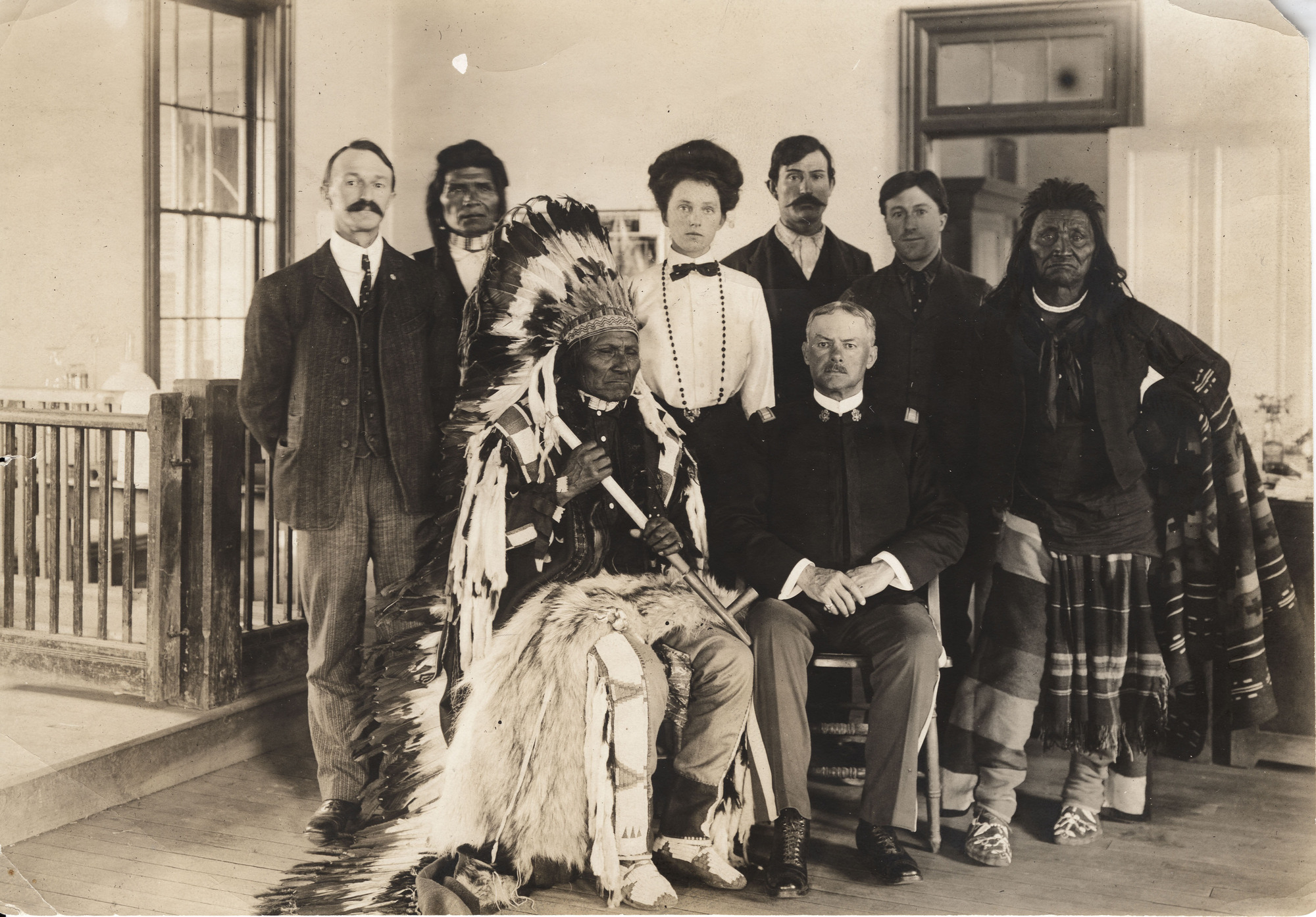 Black and white photograph of several White men and a woman posing for a photo alongside several American Indians inside a building