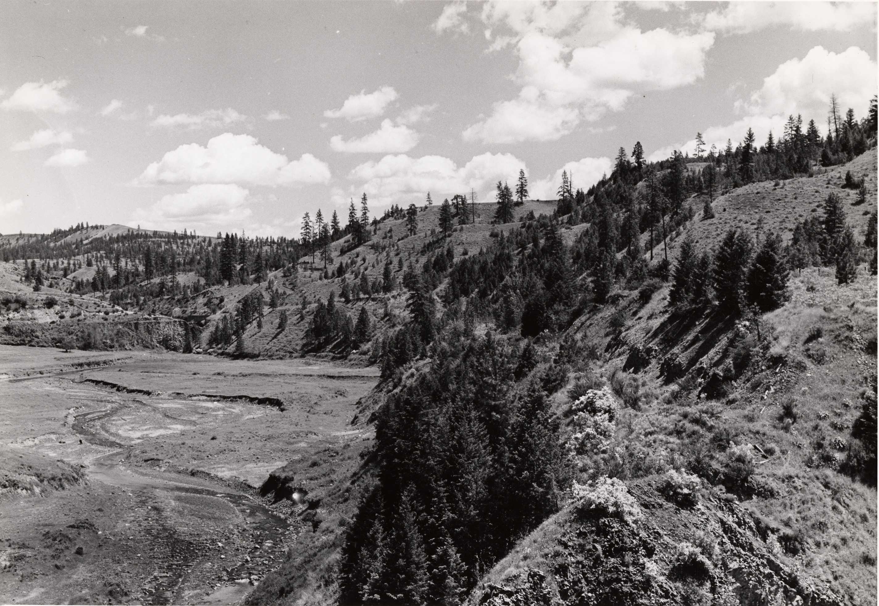 Black and white photograph of a winding creek in a valley surrounded by steep hills with scattered trees