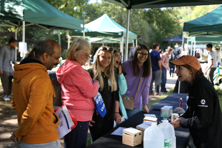 Woman in dark jacket and orange hat stands behind a table while speaking to group of smiling adults.