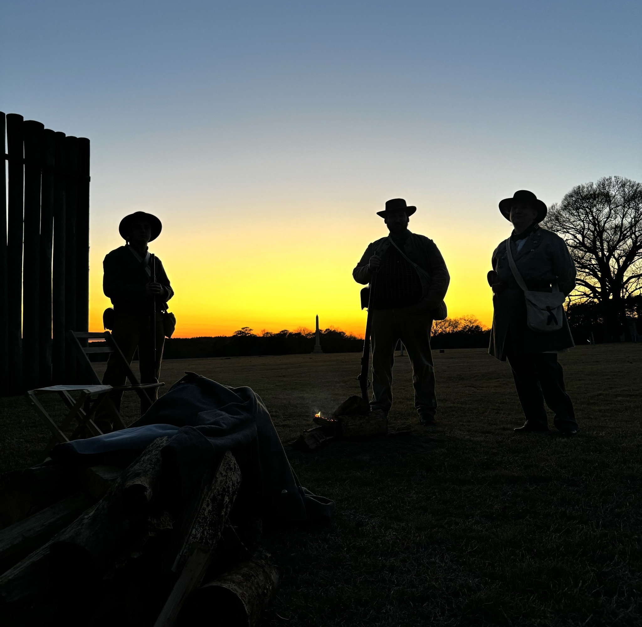 Confederate soldiers standing by a fire outside the stockade at sunset.