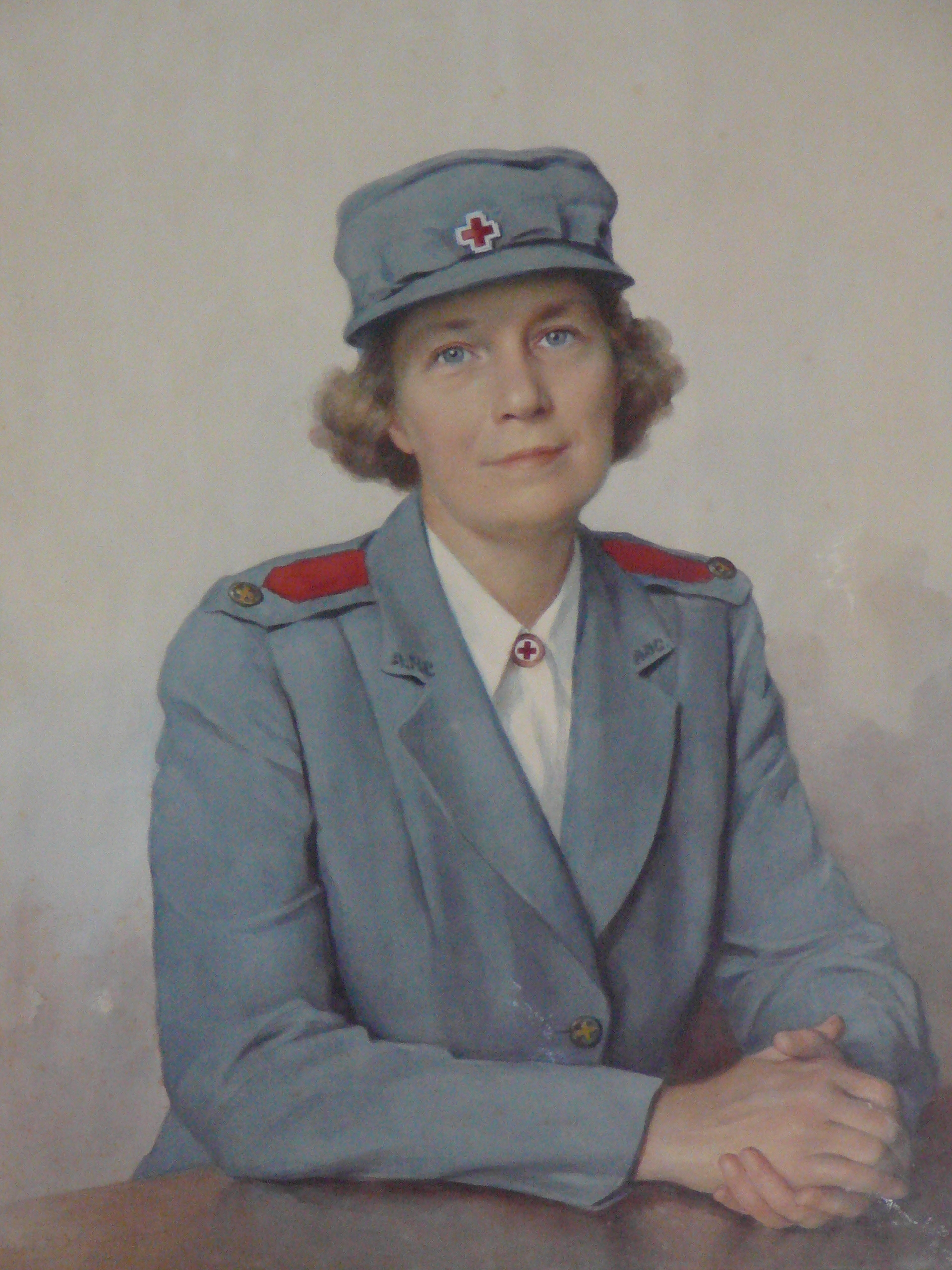 A portrait of a woman seated at a table. She is wearing a pale blue Red Cross uniform with a red cross pin on her hat and at her collar. She has short light brown hair and blue eyes. Her hands are clasped together on top of the table. She gazes at the viewer with a friendly expression. 