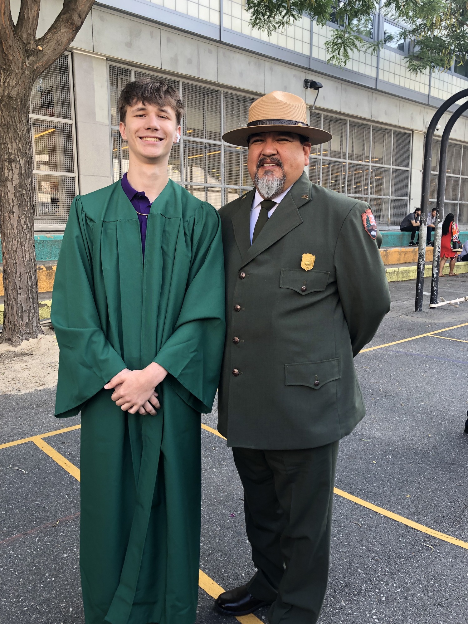 Two men stand outside smiling and posing for picture. The man on the left wears a long green graduation robe. The man on the right is wearing a class A NPS uniform and flat hat