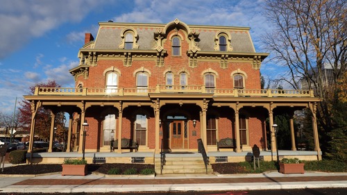 Exterior view of the Saxton home. The home is three stories with an Italianate style porch on the front. The porch consists of 8 bracketed columns and wraps around both sides of the home. The second and third story windows are arched at the top  and the roof line is decorated with red and black shingles. 