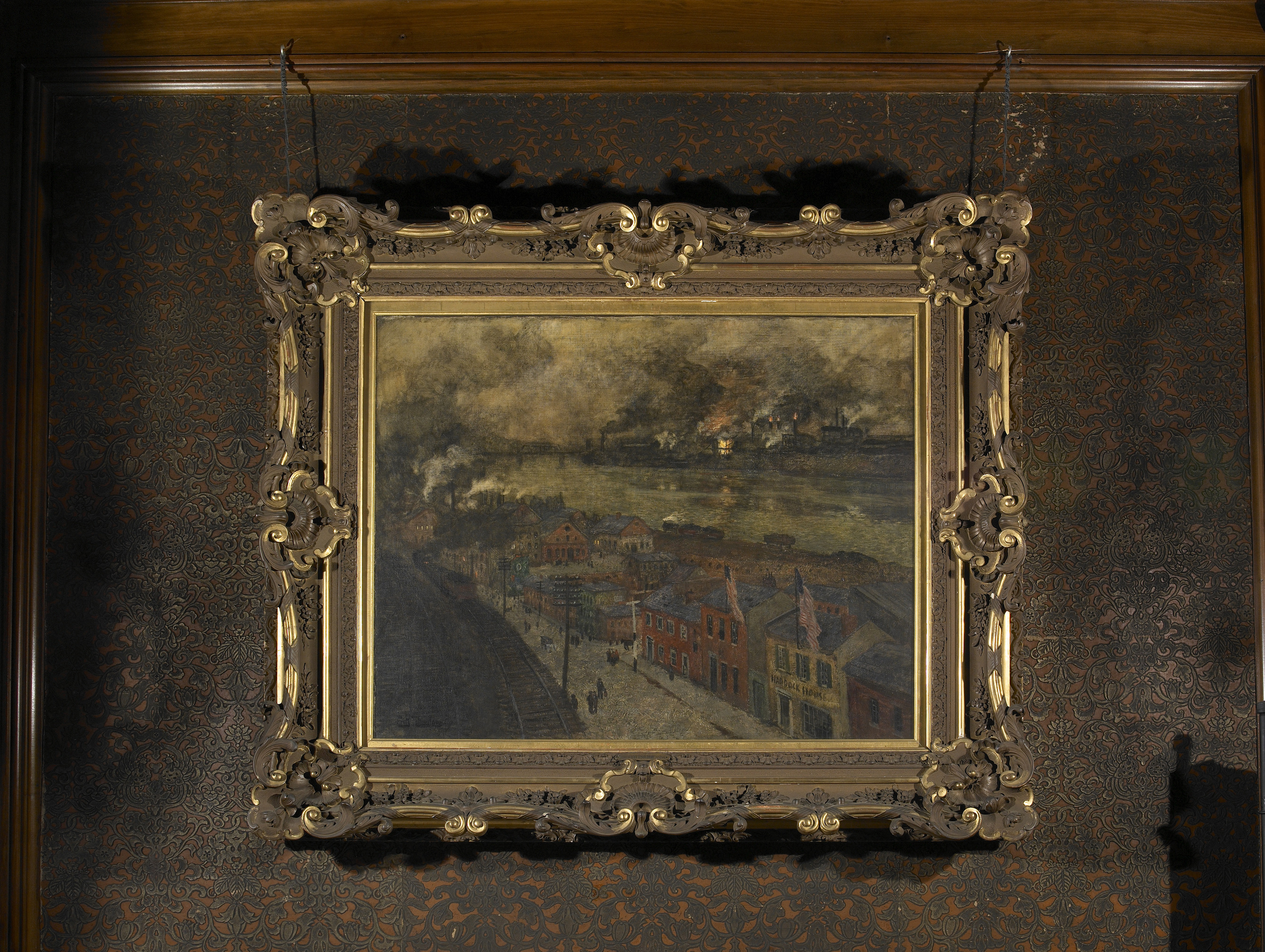 A painting that depicts a town with colorful facades of buildings being covered by smoke. There is a railroad that runs next to the town and a river that runs on the other side of the town. In the distance there are chimneys from factories that are billowing smoke and some flames burning from the smoke stacks. 