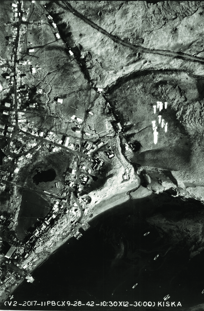 Birds-eye view of bombs dropping from U.S. plane over Kiska. Japanese ships in water below directly underneath.