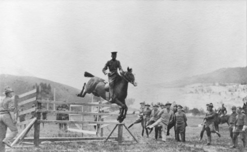 Men stand on one leg watching a horse and rider clear a jimp