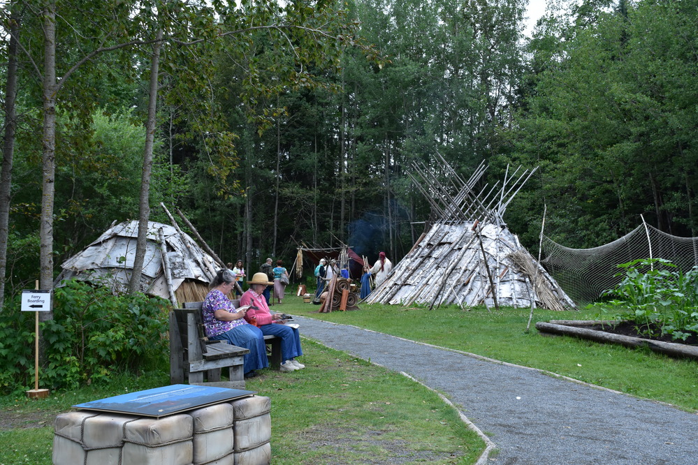 Birch bark lodges surrounded by forest with people near a path going through.