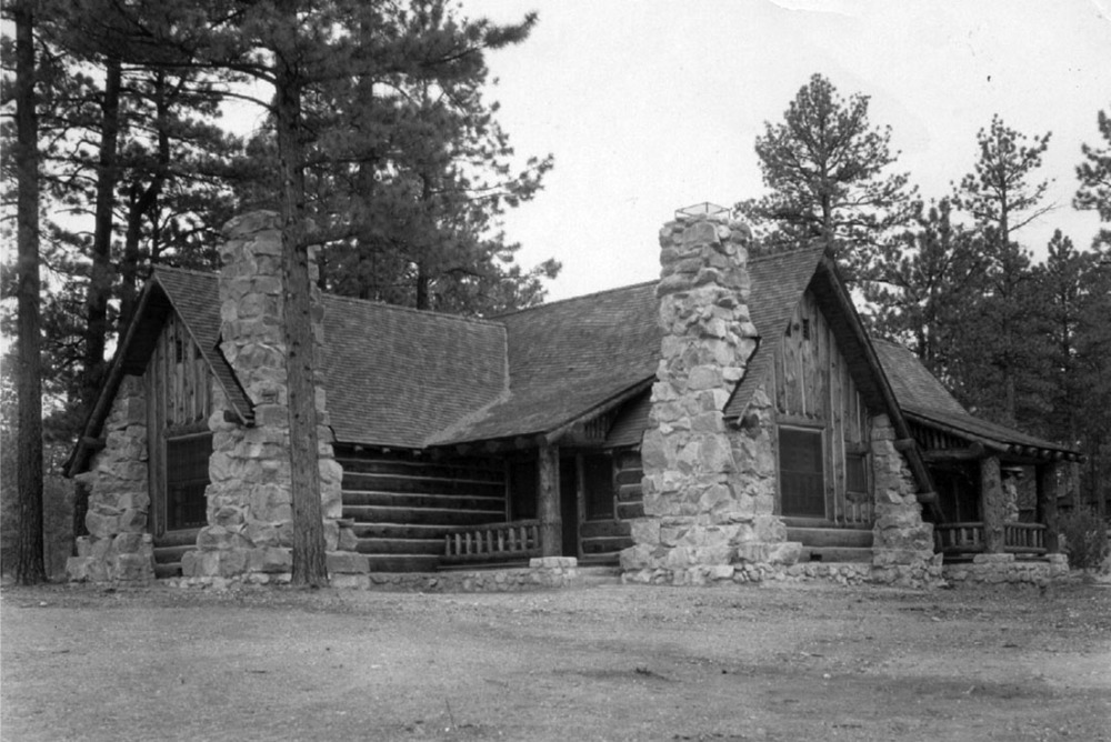 A large, squat cabin has stone pillars at the corners, a stone foundation, log walls, and a sheltered porch.
