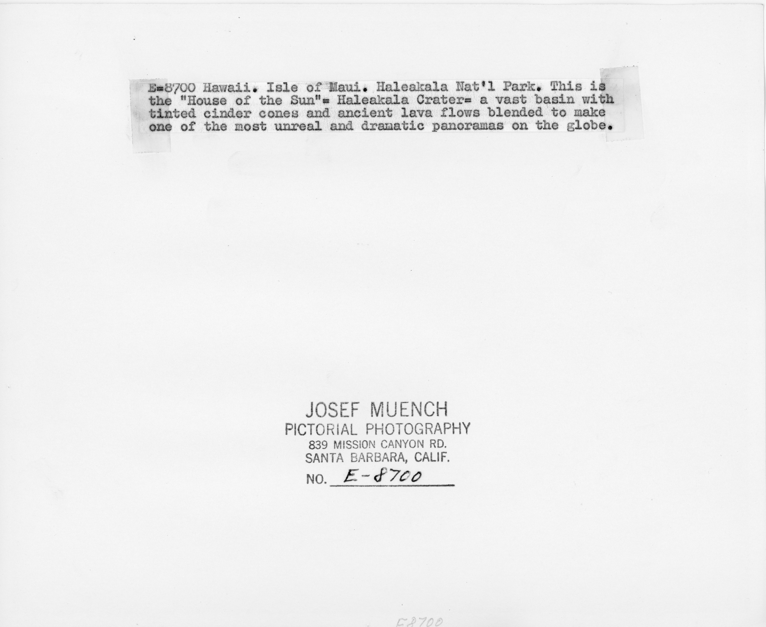 The back of the picture with mounds of cinder cones on the floor of Haleakalā crater. The back of the picture has text that reads "E-8700 Hawaii. Isle of Maui. Haleakala National Park. This is the “House of the Sun” Haleakalā Crater a vast basin with tinted cinder cones and ancient lava flows blended to back one of the most unreal and dramatic panoramas on the globe.” At the bottom, it has the photographer's name, “Josef Muench,” where the image was produced, “Pictorial Photography, 839 Mission Canyon Road. Santa Barbara, California.” Lastly, the number assigned to the image, "No. E-8700."