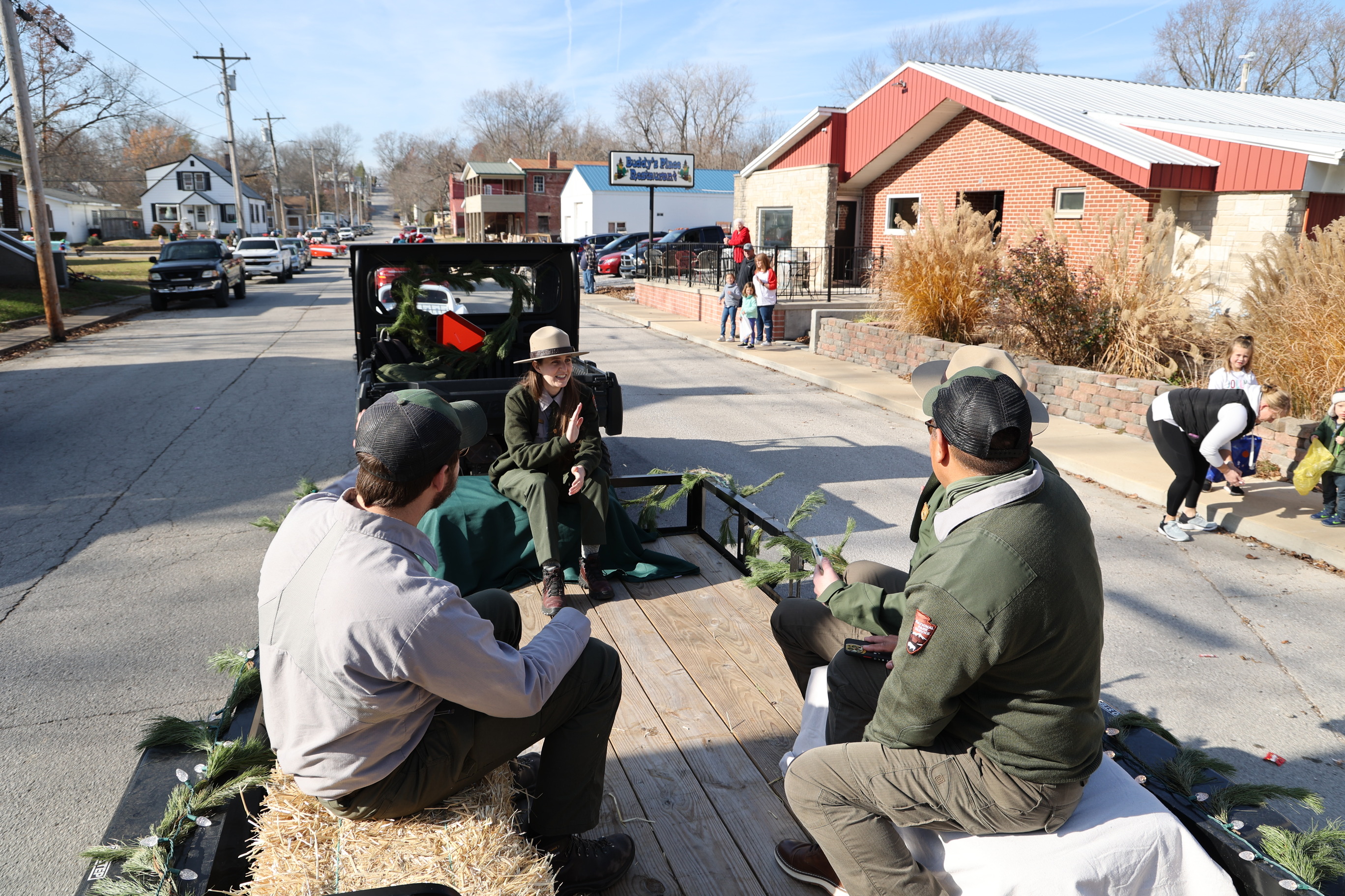 Four park staff members sit waving to people on hay bails on a wooden floored wagon decorated in green garland. 