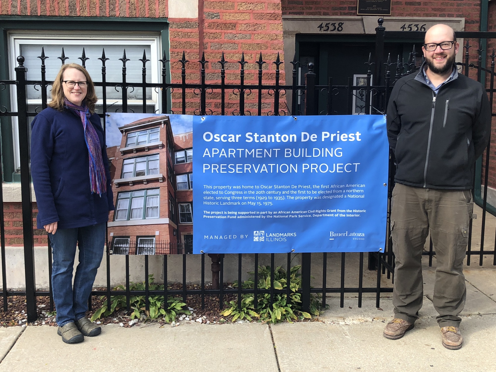 A man and a woman stand on either side of a banner outside. They are in front of a metal fence surrounding a brick building. The banner partially reads Oscar Stanton De Priest Apartment Building Preservation Project.