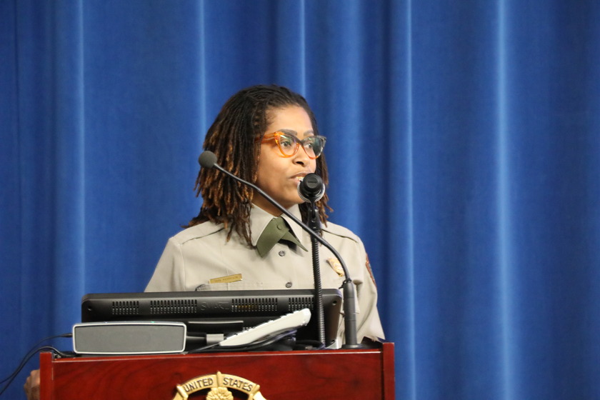 A woman with brown dreadlocks and orange-rimmed glasses stands at a dark wooden podium. She wears a short-sleeved NPS shirt with a dark tie. Her head is turned slightly to the left as she looks forward. A microphone stands in front of her. In the background is a dark blue curtain.