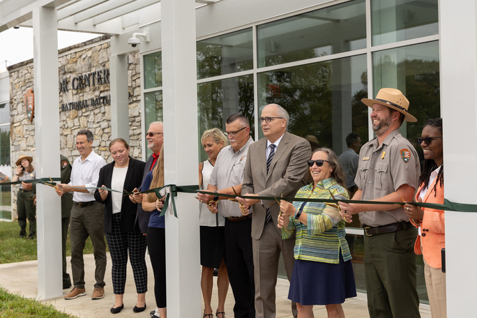 A group of park rangers and partners cut a green ribbon attached to columns in front of a large visitor center