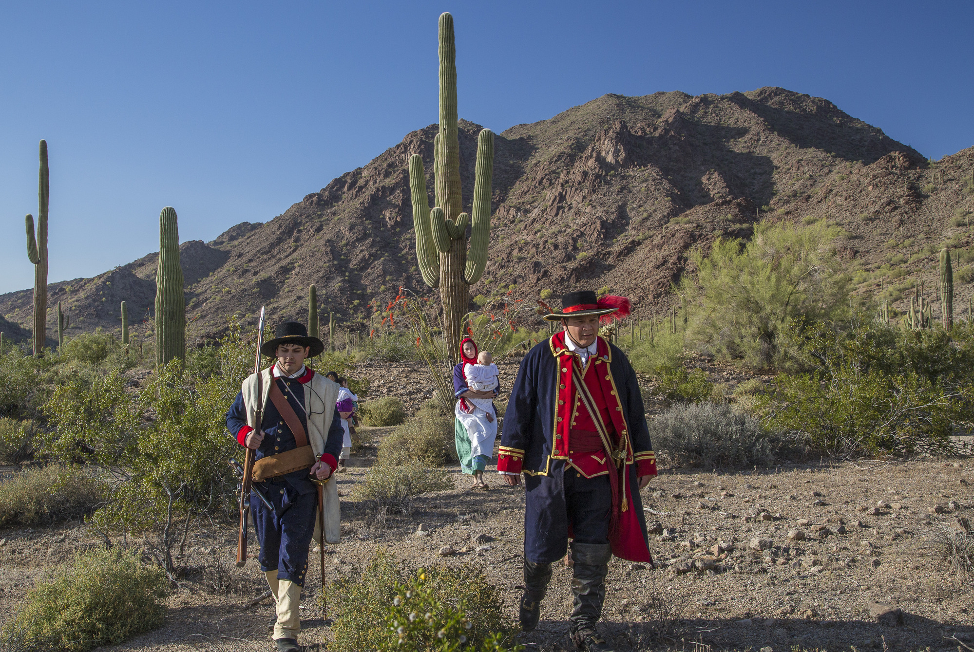 Two men wearing Spanish colonial military uniform and two women carrying children walk through the desert