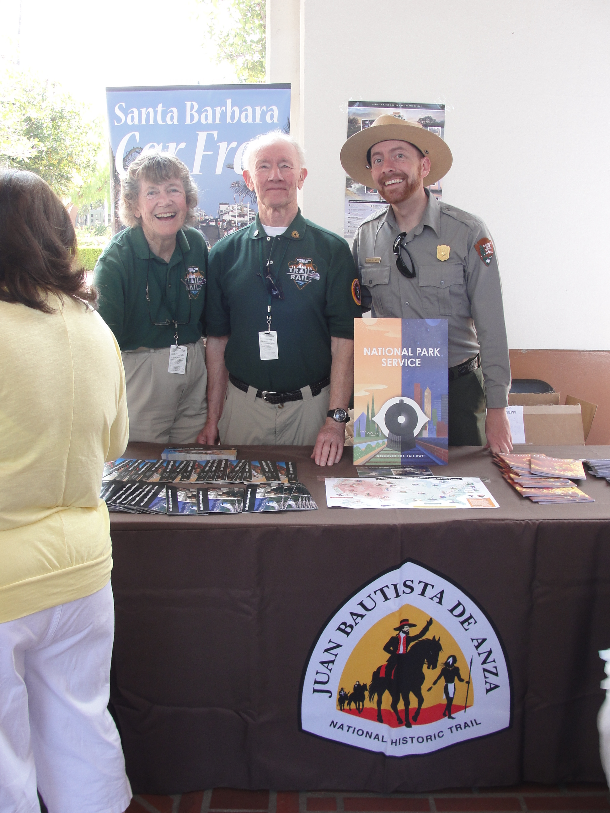 Two NPS volunteers wearing green shirts stand next to each other and an NPS ranger smiling behind an Anza Trail table