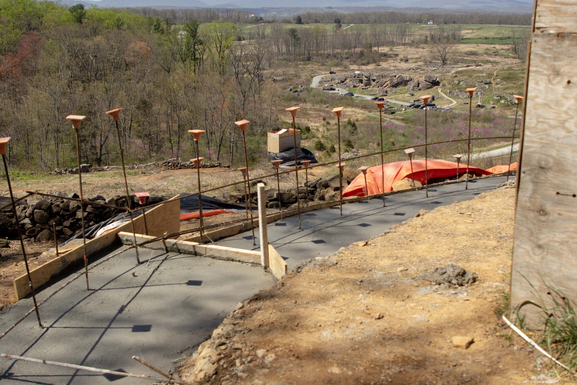 A concrete pad with boards and metal construction stakes runs alongside a monument in the foreground, with rocks, trees, and monuments in the background.