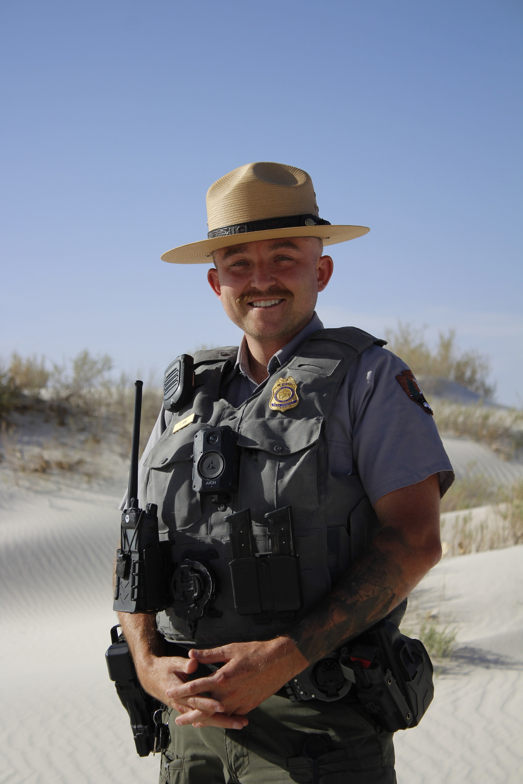 A man in a law enforcement park ranger uniform stands for a photo. White sand dunes and desert shrubbery under a pale blue sky fill his surroundings.  
