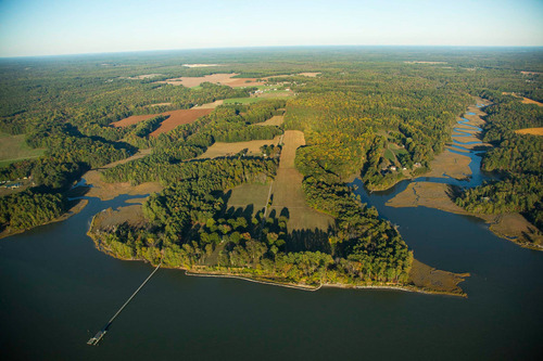 Aerial view of a river shoreline with fields and forest stretching out into the distance. A pier is visible in the foreground. 