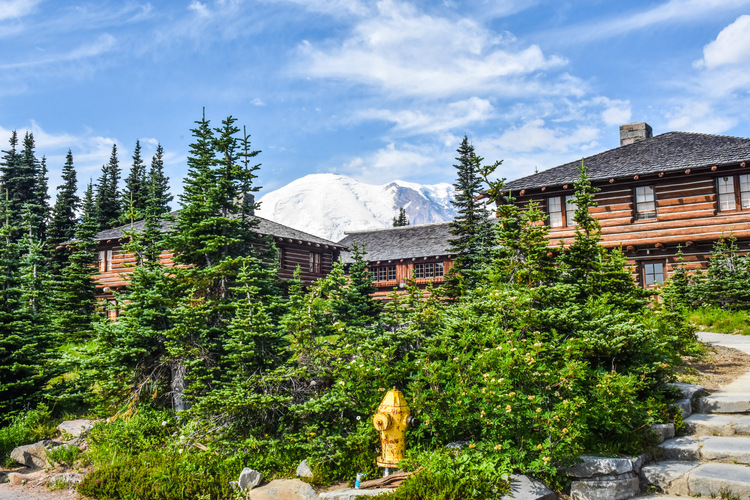 A wooden, rustic building sits in front of a glaciated mountain peak, surrounded by green trees.