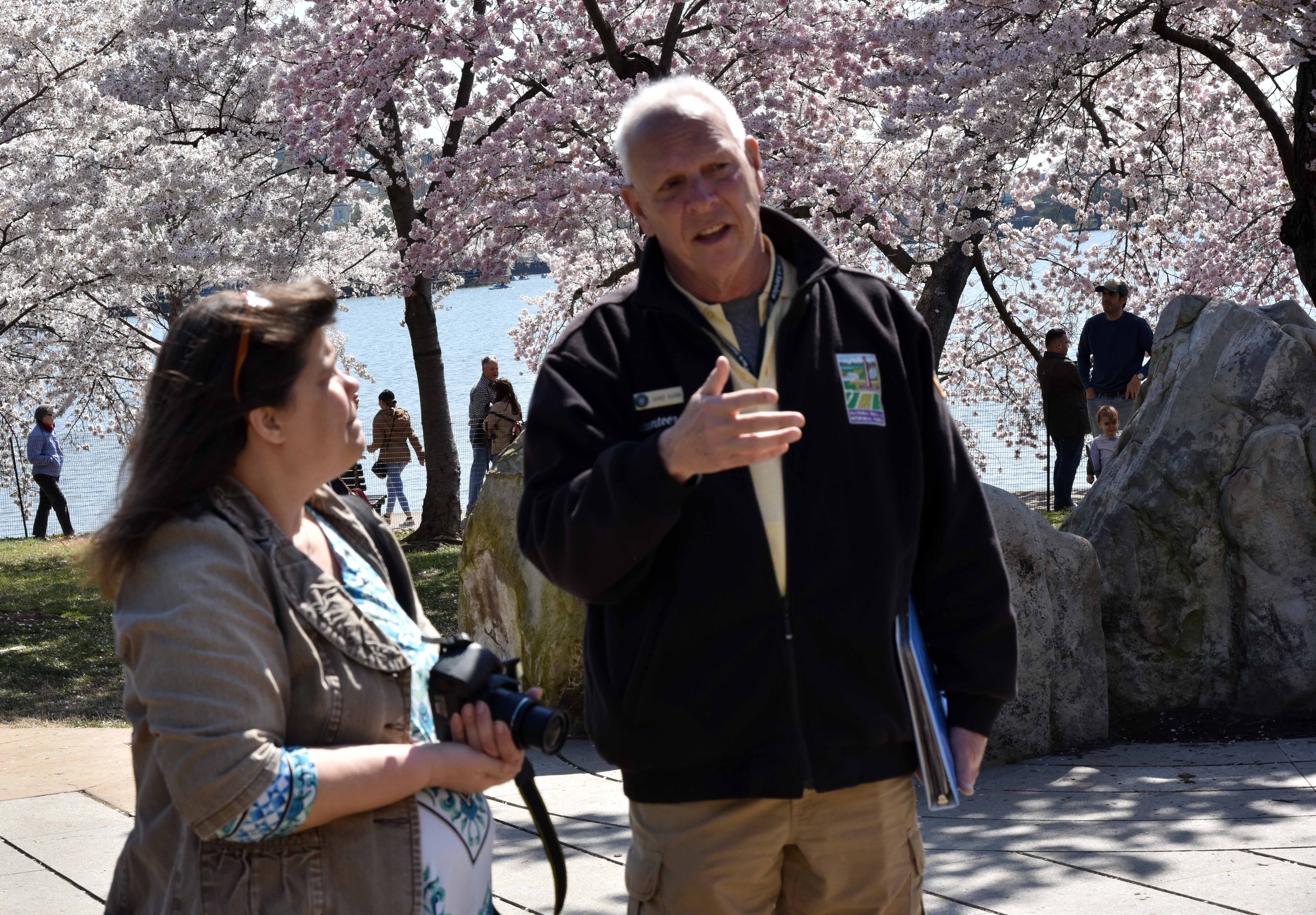 Man talks to a woman holding a camera with blooming cherry trees in background.