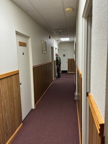 View down a long hallway with wood paneling and purple carpeting. Man in green jacket stands at the far end of the hallway. 