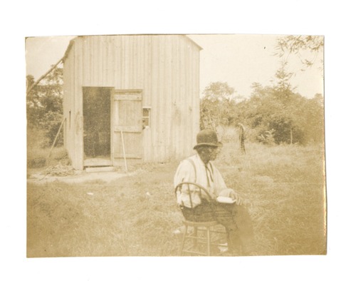 An older gentleman wearing a round top hat sits on a chair outside of a wooden house