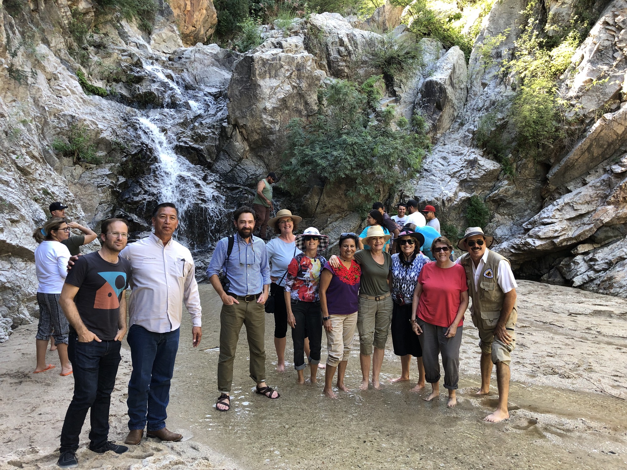A group of people stand in a shallow pool in front of a waterfall