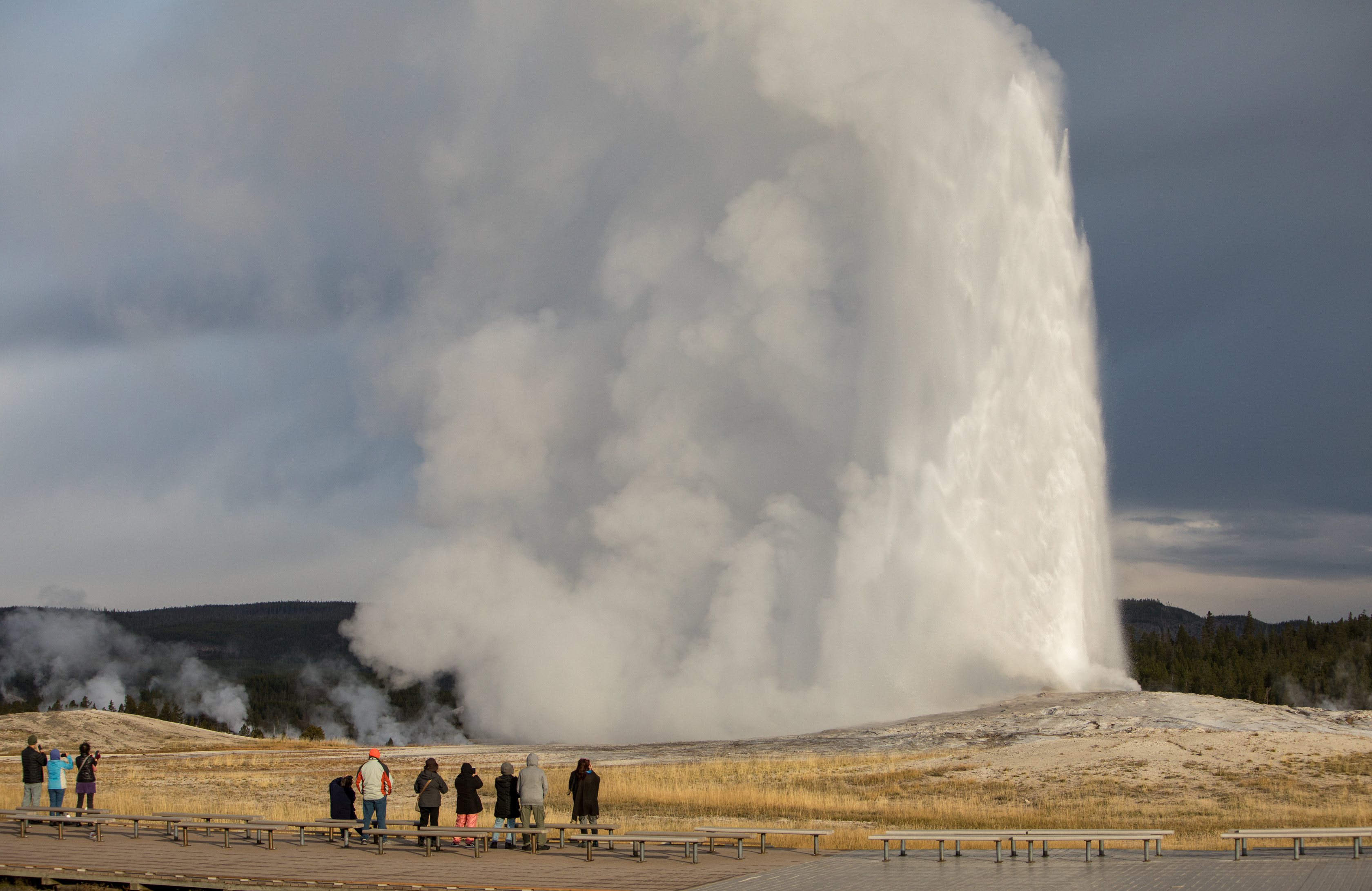 A geyser eruption with visitors standing on the boardwalk watching.