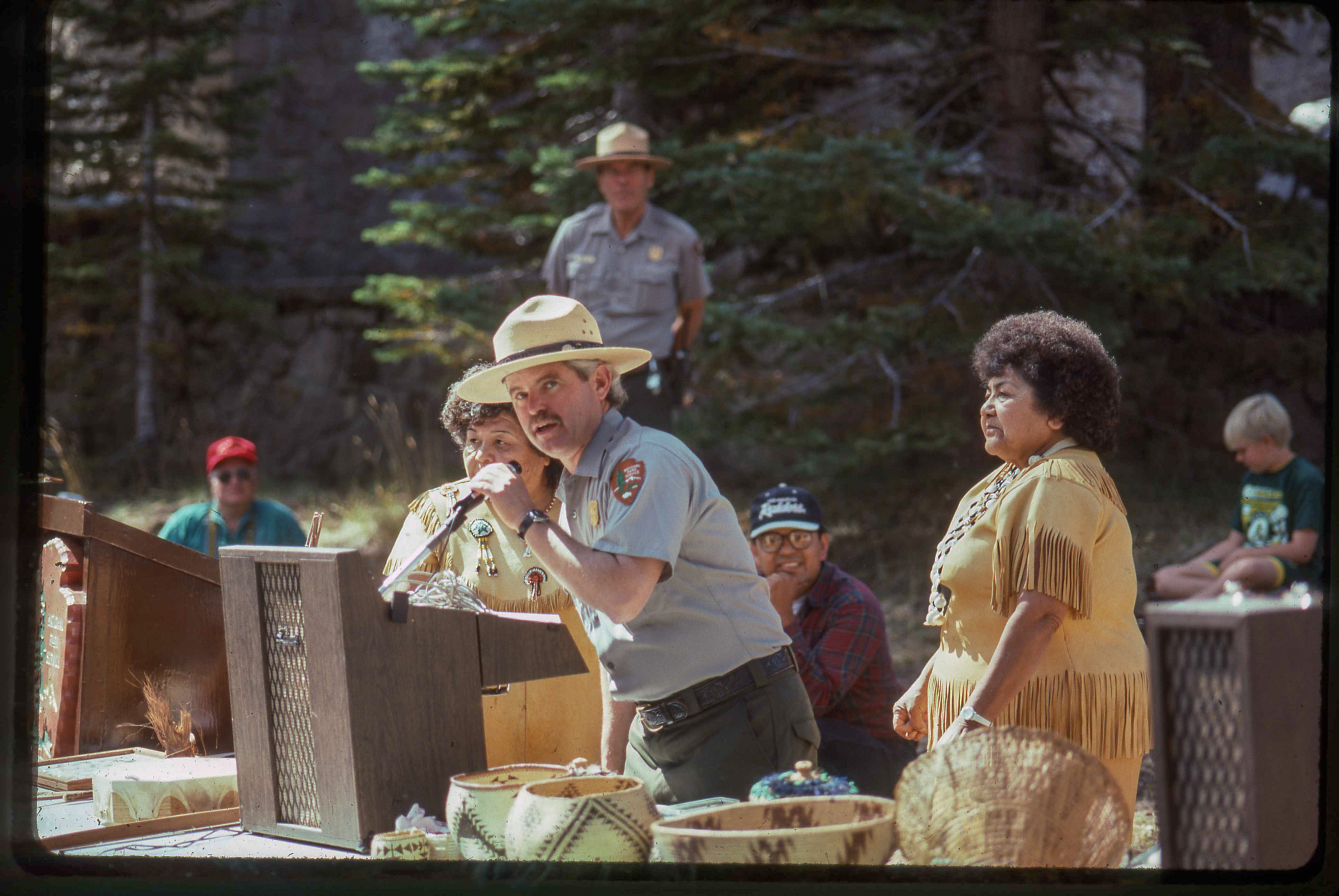 A man speaks at a podium flanked by two women in traditional American Indian attire at an event in the Manzanita Lake Area.